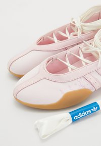 Sapatilhas de ballet em lona rosa com um detalhe em couro, design de atilhos abertos e sola em borracha, acompanhadas de uma fita branca e um rótulo azul com marca.