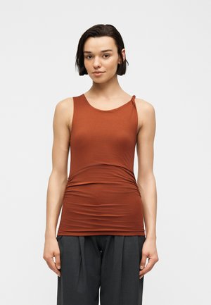 Young woman with short dark hair wearing a rust-colored sleeveless top and dark gray loose pants, standing against a light background.