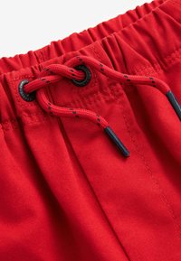 Shorts en tissu rouge avec ceinture élastique, cordon de serrage ajustable de couleur contrastante, et embouts en plastique noir. Texture lisse avec détail de couture.