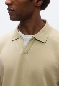 Bézs színű galléros póló V-nyak kialakítással és texturált anyagból, fehér kerek nyakú inggel rétegezve. Közeli kép a gallérról és a nyakkivágásról.