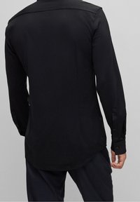 Maglia nera a maniche lunghe con taglio dritto, texture liscia e polsini con bottoni. La parte posteriore presenta un dettaglio sottile a yoke e si estende leggermente più a lungo.