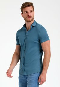 Polo majica s kratkimi rokavi v enotni teal barvi, izdelana iz mehkega materiala, s knežno zapenjanjem in diskretno oznako na prsih.