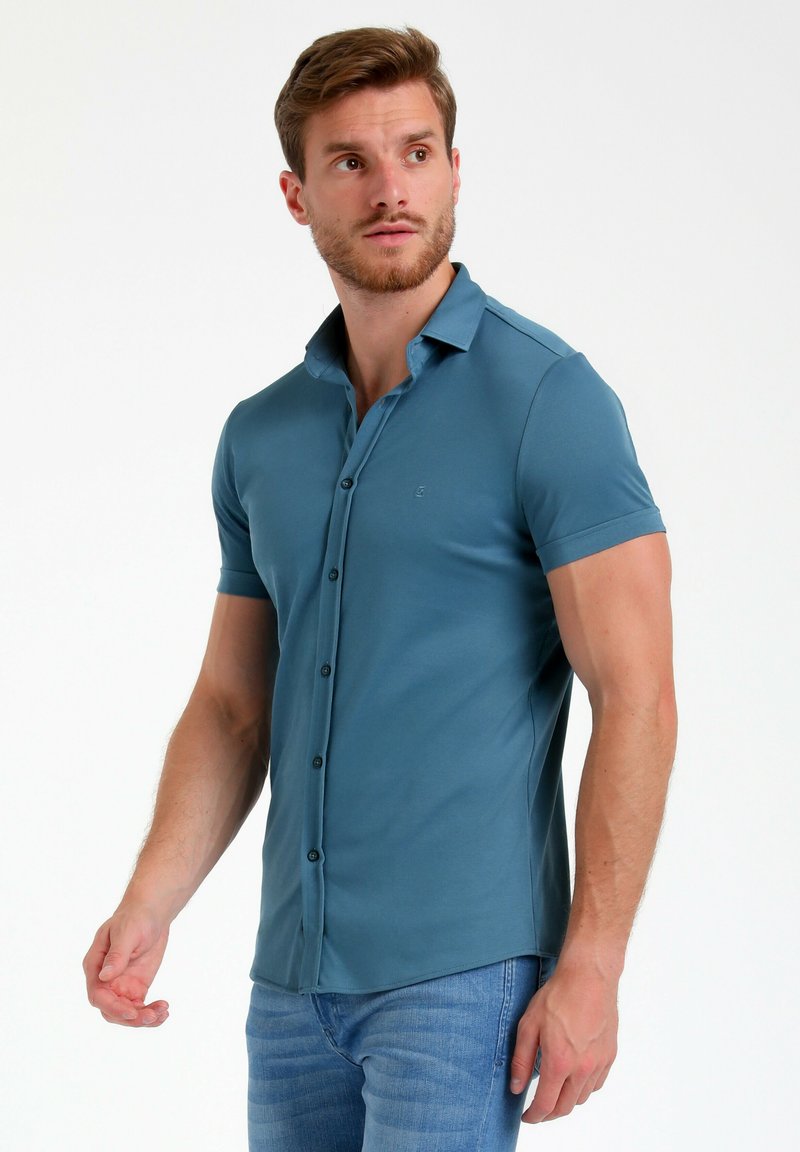 Polo majica s kratkimi rokavi v enotni teal barvi, izdelana iz mehkega materiala, s knežno zapenjanjem in diskretno oznako na prsih.