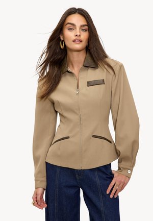 Vrouw in een beige jack met ritssluiting, voorzien van een donkerbruine kraag en zakaccenten, gecombineerd met donkerblauwe jeans, poseert met haar hand op de heup.