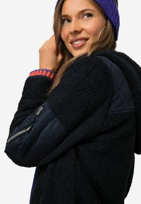 Giacca in pile nera con maniche sintetiche trapuntate. Presenta polsini a coste arancioni e viola. Tasca con zip sul braccio sinistro, tessuto testurizzato.