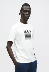 Jonge man met dreadlocks, gekleed in een wit T-shirt met de tekst "BOSS" en een verloopontwerp, en een blauwe broek, staand met zijn handen in zijn zakken.