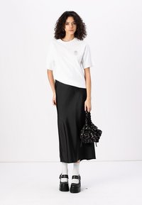 T-shirt blanc à manches courtes avec un logo, associé à une jupe midi en satin noir, des chaussettes blanches, des grosses chaussures à plateforme noires et un sac à motifs.