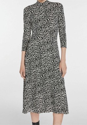 Vrouw draagt een zwart-witte jurk met een abstract patroon, tot aan de knie, met driekwart mouwen en een hoge halslijn, staand tegen een effen achtergrond.