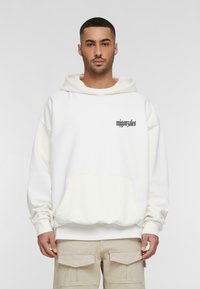 Sudadera blanca con capucha y un bolsillo frontal. Diseño minimalista, ajuste ligeramente oversize, con un logo negro en el pecho. Textura suave.