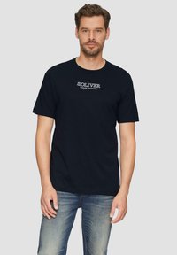Marineblaue Baumwoll-T-Shirt mit kurzen Ärmeln, Rundhalsausschnitt und weißem "S. OLIVER CASUAL APPAREL"-Logo auf der Brust. Getragen mit hellblauen Jeans.