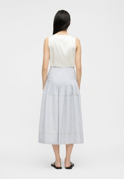 Femme debout de dos, portant un haut blanc sans manches, une jupe midi taille haute à rayures verticales et des sandales plates noires.