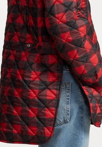 Giacca trapuntata in plaid rosso e nero, con chiusura a pressione e fessure laterali, abbinata a denim azzurro chiaro. Materiale leggero e strutturato.