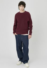 Pull en tricot côtelé bordeaux associé à un jean bleu foncé à coupe décontractée et des baskets blanches avec des accents gris et verts.