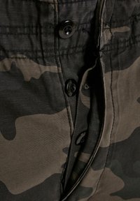 Pantalones cortos de camuflaje con un patrón de camuflaje en negro y verde, cierre con botón y tela suave y texturizada. Primer plano del detalle de la cinturilla.