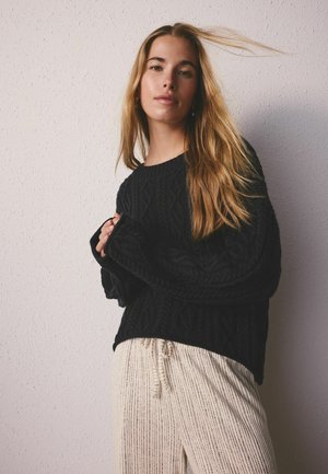 Ung kvinde med langt blondt hår iført sort kabelstrikket sweater og cremefarvede ribstrikkede bukser, stående mod en lys struktureret væg.