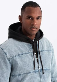 Light blue denim jacket with a black hooded sweatshirt underneath. The jacket features a front zipper and contrasting stitching.