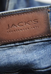 Denim w jasnoniebieskim odcieniu, z brązową skórzaną metką wytłoczoną napisem "JACK'S SPORTSWEAR INTL," przymocowaną widocznym szwem.