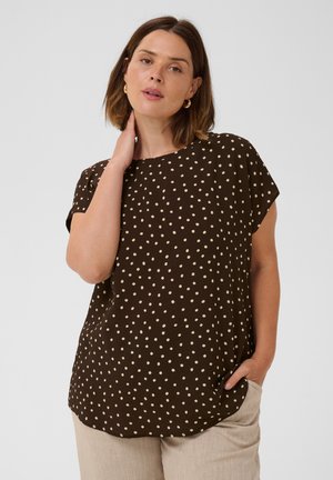 Femme aux cheveux bruns mi-longs portant un chemisier marron à manches courtes avec des pois clairs et un pantalon beige, main sur le cou et dans la poche.