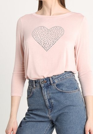 Femme portant une chemise rose clair à manches longues avec un motif de cœur clouté, associée à un jean bleu taille haute.