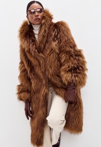 Brun kunstpelsjakke med en stor krave, lange ærmer og tekstureret finish. Stylised med beige bukser og brune læderhandsker.