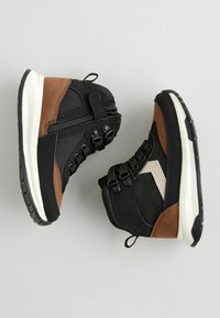 Bottes de sport noires et brunes avec une tige en tissu texturé, dotées d'une fermeture éclair sur le côté, d'un devant à lacets et de semelles intermédiaires blanches, inclinées pour l'affichage.
