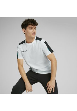 T-shirt sportivo bianco con accenti neri sulle spalle, logo sul petto e dettagli teal. Il modello indossa pantaloni neri; sfondo neutro.