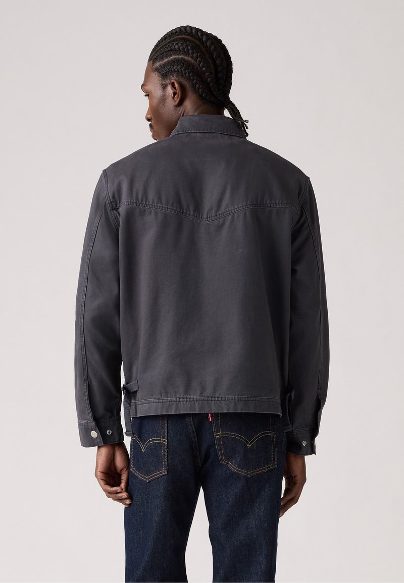 Graue Canvas-Jacke mit strukturiertem Design, Druckknöpfen und taillierter Passform, mit einer Schulternahtdetails auf der Rückseite und seitlichen Laschen.