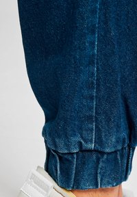 Joggers de mezclilla en azul oscuro con una tela texturizada, que presentan puños elásticos y detalles de costura visibles. Llevados con zapatillas blancas.