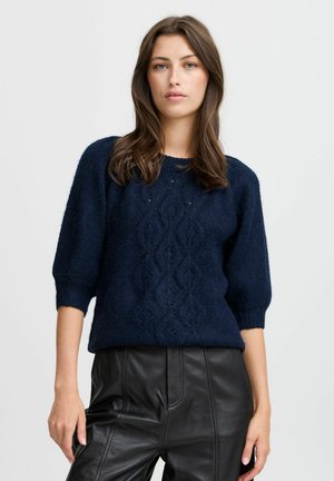 Marineblauer Pullover mit strukturiertem Zopfmuster, Rundhalsausschnitt und kurzen Ärmeln. Getragen mit schwarzen Lederhosen.