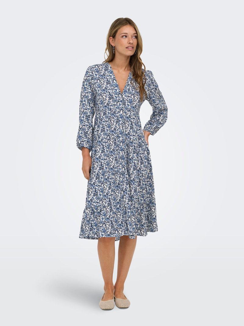 Femme debout dans une robe midi fleurie bleu et blanc à manches longues, col en V, et chaussures plates beiges sur un fond clair uni.