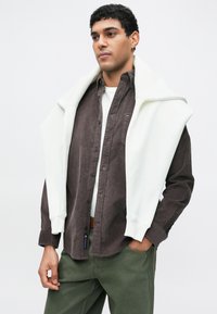 Ανδρικό σύνολο με καφέ πουκάμισο κοτλέ με κουμπιά και λευκό φούτερ που είναι περασμένο στους ώμους. Παντελόνι σε ελιάς πράσινο χρώμα. Εμφανές λογότυπο γνωστής μάρκας στο πουκάμισο.