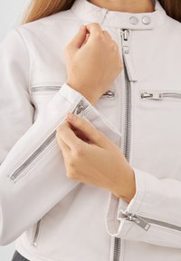 Vit läderjacka med hög krage och silverdragkedjor. Har två dragkedjefickor på bröstet och dragkedjor vid ärmsluten för justerbarhet.