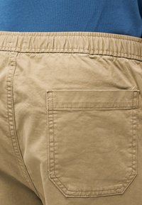 Κοντινή λήψη τσέπης παντελονιού με ελαστική μπεζ μέση και λεπτομερή ραφή, με φόντο μπλε πουκάμισο.