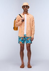 Camisa de manga larga naranja con estampado, camiseta blanca, pantalones cortos azules con diseño de peces, zapatos marrones tipo slip-on, bolsa amarilla impermeable, sombrero de cubo, gafas de sol.