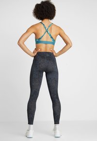 Sportbh i turkos med korsande baksidesdesign, ihop med mörka, mönstrade leggings. Materialet verkar stretchigt och åtsittande.