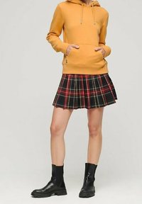 Žlutá mikina s kapucí a přední kapsou, spárovaná s černou a červenou kostkovanou skládací sukni. Černé kotníkové boty dotvářejí outfit.