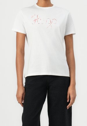 Persona con una camiseta blanca de manga corta con "Hugo" bordado en letra rosa, combinada con pantalones negros sobre un fondo liso.