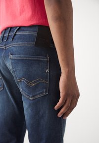 Mörkblå jeans i denim med guldstickningar på bakfickorna, utrustade med en svart läderetikett vid midjan. En figurnära design.