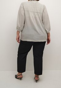 Blouse rayée en crème clair et gris foncé, avec un dos froncé et des manches bouffantes. Associée à un pantalon noir bien taillé et des talons.