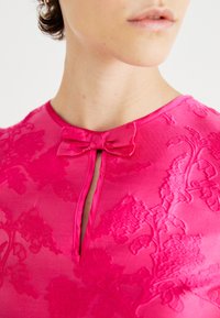 Fuchsia top with a floral embossed pattern, featuring a keyhole neck and a bow accent at the collar. Smooth texture and fitted design.