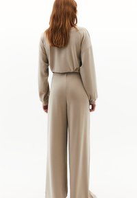 Mono de color beige claro con un ajuste relajado, que presenta mangas largas y piernas anchas. Tejido suave con un detalle ligeramente fruncido en la parte posterior.