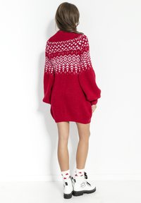 Kobieta ubrana w czerwoną, dużą dzianinową sukienkę z białymi geometrycznymi wzorami na górze, białe skarpetki z czerwonymi gwiazdkami oraz białe botki na sznurowadła.