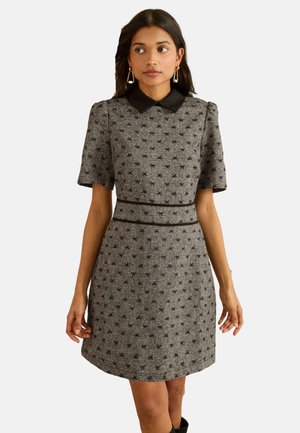 Woman wearing a grey dress with black bow patterns, black collar, short sleeves, and black piping at the waist, standing with hands relaxed.