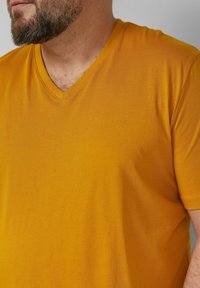 Geel T-shirt met V-hals van zacht materiaal, met een minimalistisch ontwerp en een subtiel logo in de linkerbenedenhoek, korte mouwen.