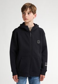 Sudadera con cremallera negra de tejido grueso con un bolsillo frontal, capucha ajustable y un parche con el logo en el pecho, con detalles de texto en la manga.