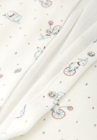 Tela blanca con pequeñas ilustraciones bordadas de conejos y triciclos en azul y rojo, que cuenta con botones de presión plateados a lo largo de la tapeta.