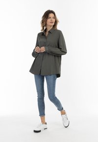 Camicia button-up verde scuro con maniche lunghe e polsini arricciati, abbinata a jeans skinny blu chiaro e sneakers bianche.