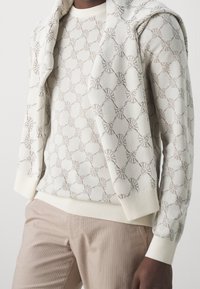 Strickpullover in Creme mit geometrischem Muster, kombiniert mit einem passenden leichten Schal. Creme- und Beigetöne, gerippte Bündchen und Saum.