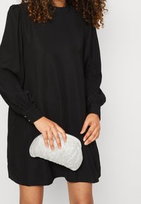 Abito nero a maniche lunghe con dettagli arricciati sulle spalle, abbinato a una borsa a tracolla con strass argentati tenuta in una mano.