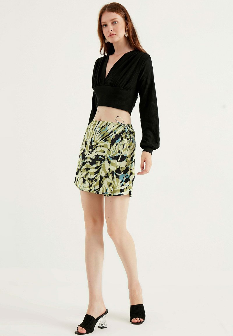 Zwarte crop top met een diepe V-hals, gecombineerd met een groene en zwarte abstracte minirok. Voorzien van open-toe zwarte hakken.